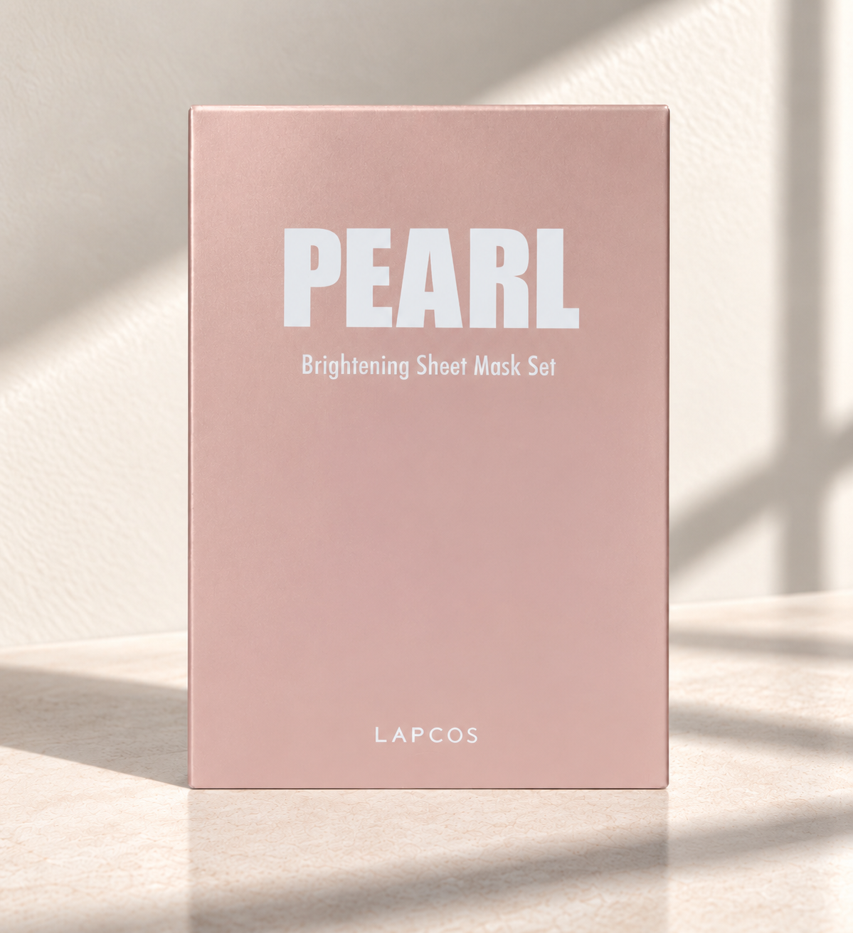 A tall rectangular pink box labeled 'PEARL Brightening Sheet Mask Set' with 'LAPCOS' at the bottom, standing on a light surface with soft window shadows.