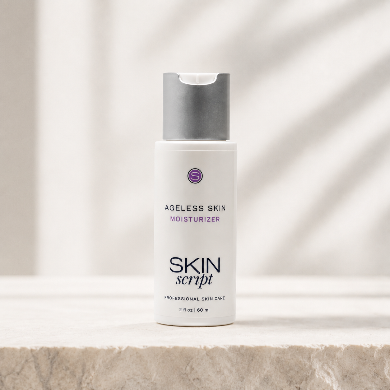 A white bottle of Skin Script Ageless Skin Moisturizer with a gray cap, standing on a light stone surface against a soft white background with leaf shadows.