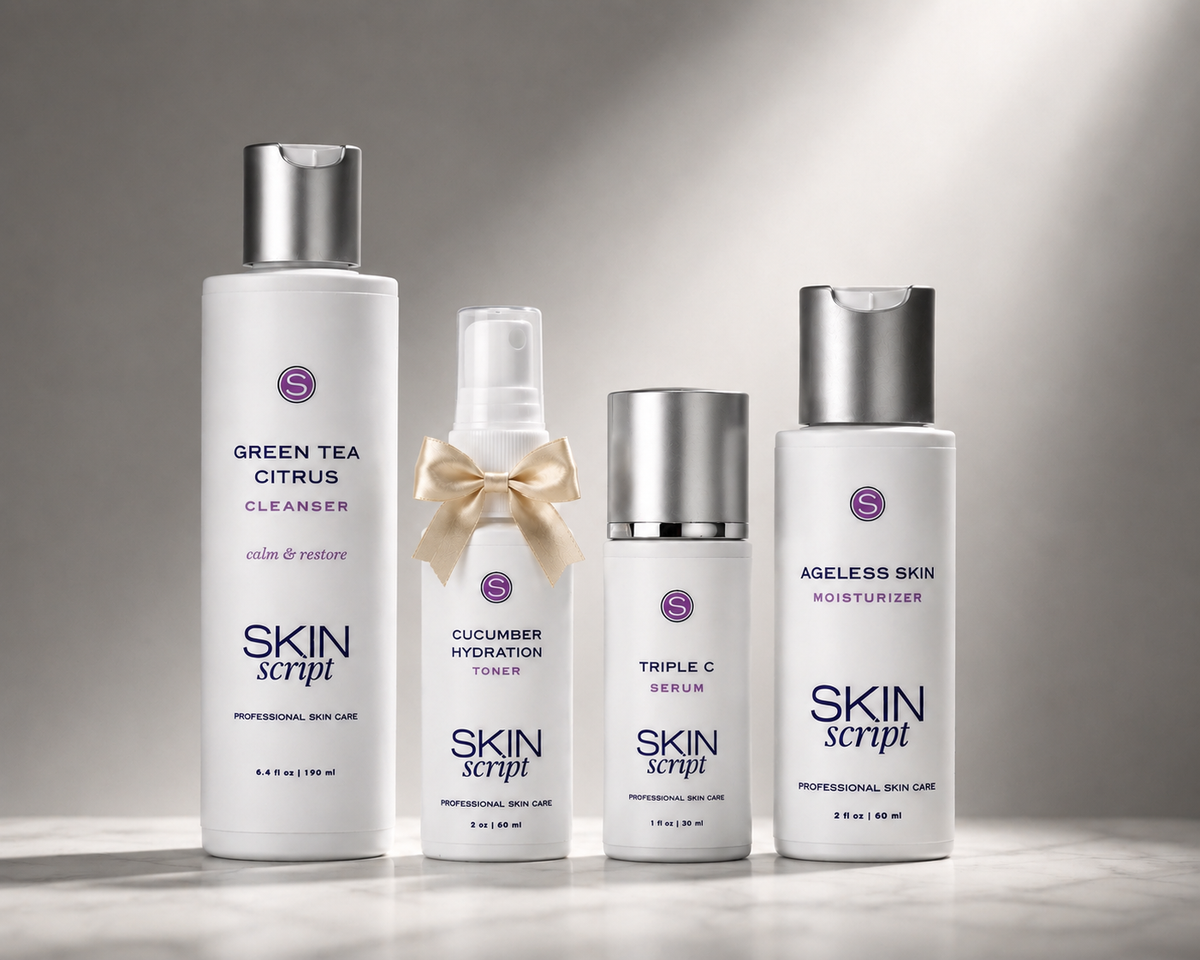 Four SKIN script skincare bottles arranged on a light gray surface against a soft gray background. The set includes a Green Tea Citrus Cleanser bottle, a Cucumber Hydration spray with a cream bow, a Triple C Serum bottle, and an Ageless Skin Moisturizer.