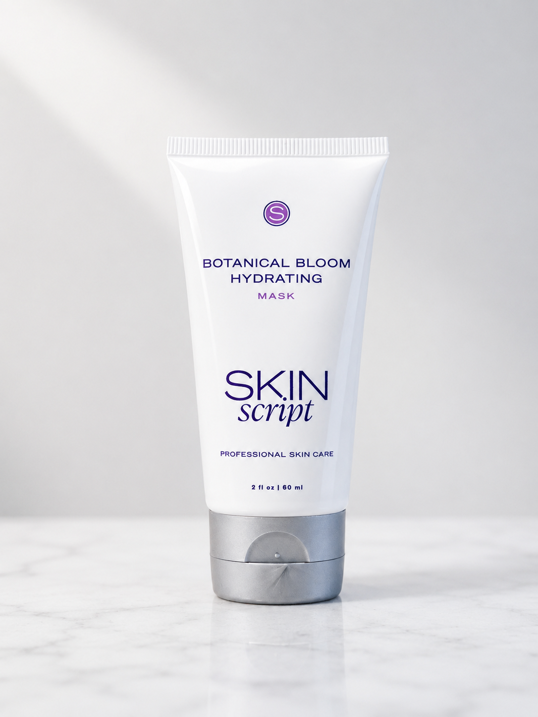 A white squeeze tube of Skin Script Botanical Bloom Hydrating Mask with a silver cap, standing on a light marble surface against a soft gray background.