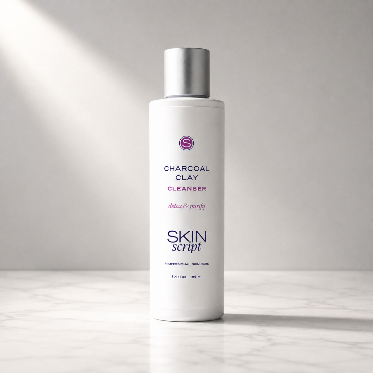A white bottle of Skin Script Charcoal Clay Cleanser with a metallic silver cap, standing on a light marble surface against a gray background with soft diagonal light rays.
