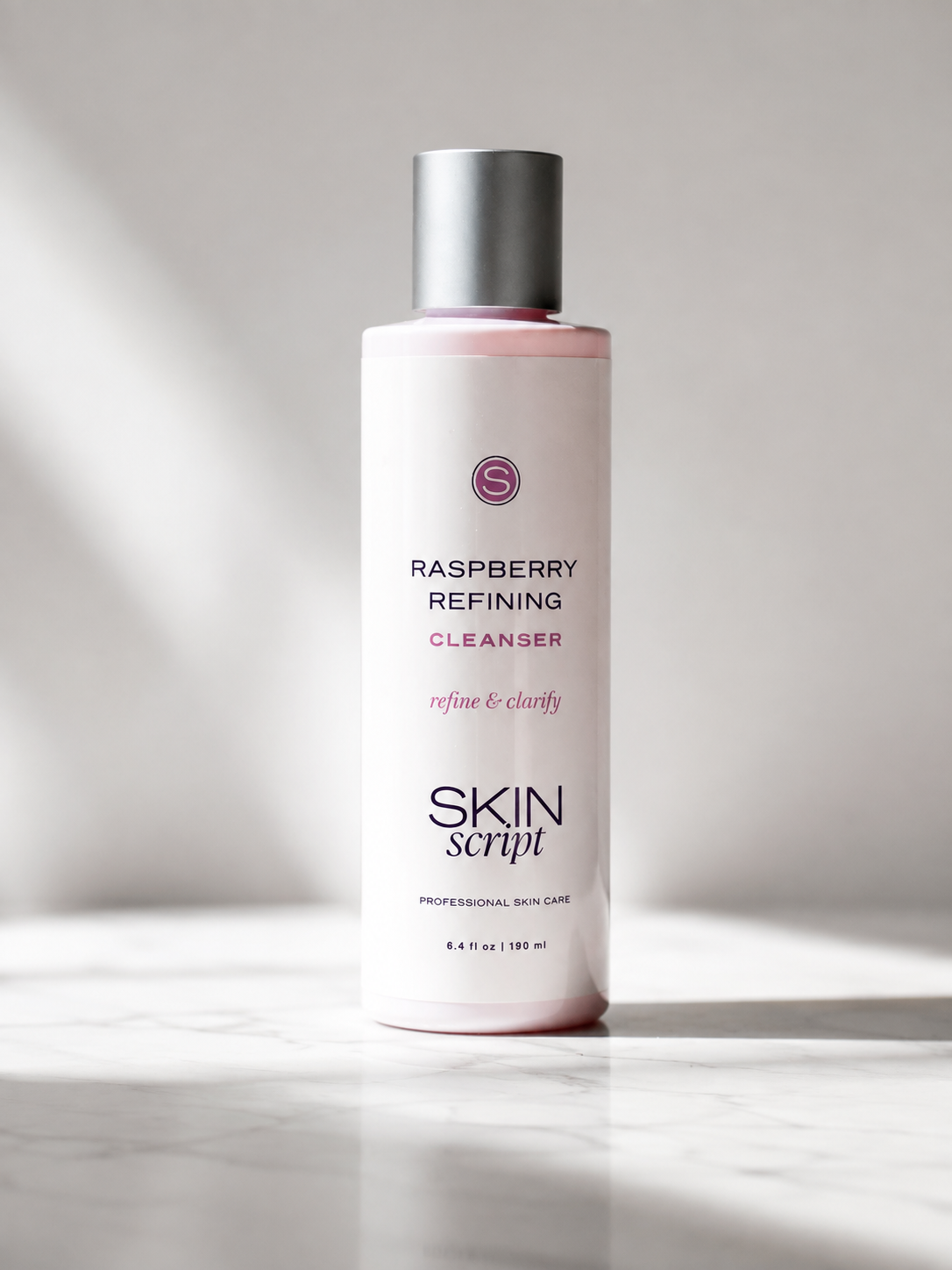 A bottle of Skin Script Raspberry Refining Cleanser with a silver cap and a pale pink label, set on a marble surface with soft diagonal shadows.