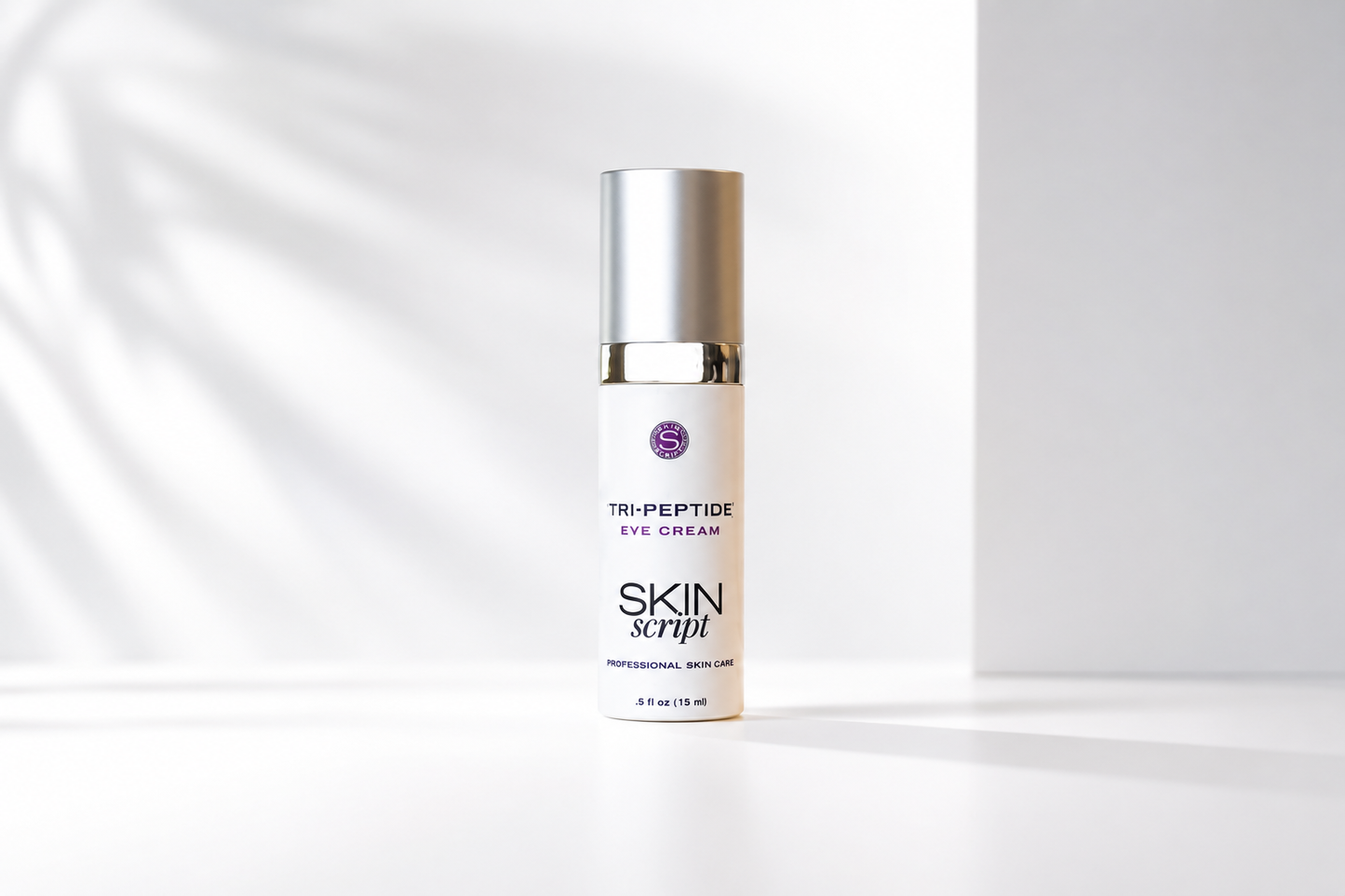 A bottle of Skin Script Tri-Peptide Eye Cream with a silver cap and white bottle, standing on a white surface with soft leaf shadows on a light gray background.