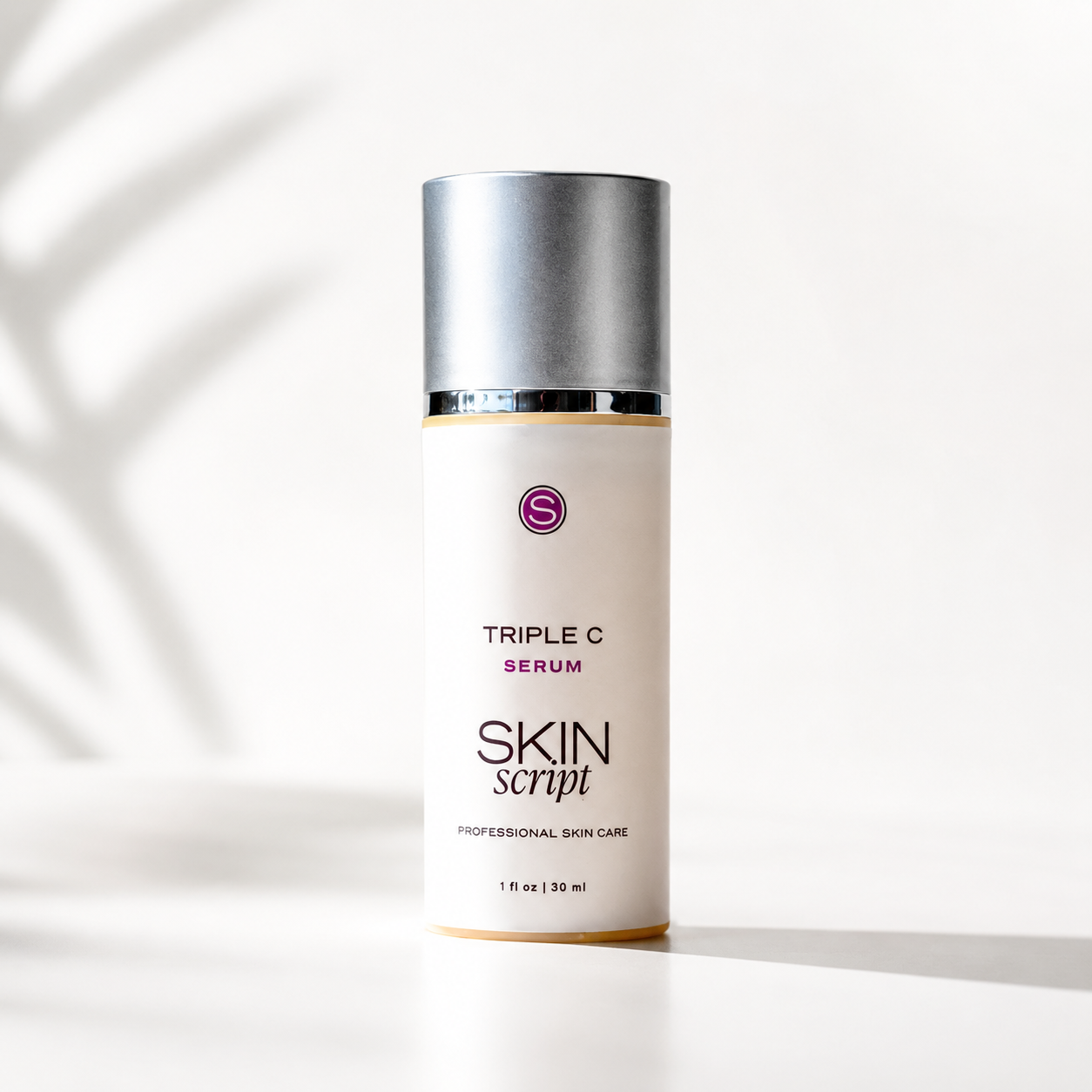 A bottle of Skin Script Triple C Serum with a white label and metallic silver cap, standing on a white surface with soft leaf shadows cast across it.
