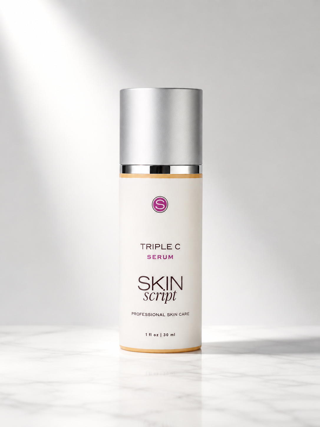 A bottle of Skin Script Triple C Serum with a white body, silver cap, and gold accents, standing on a light marble surface against a soft gray background.